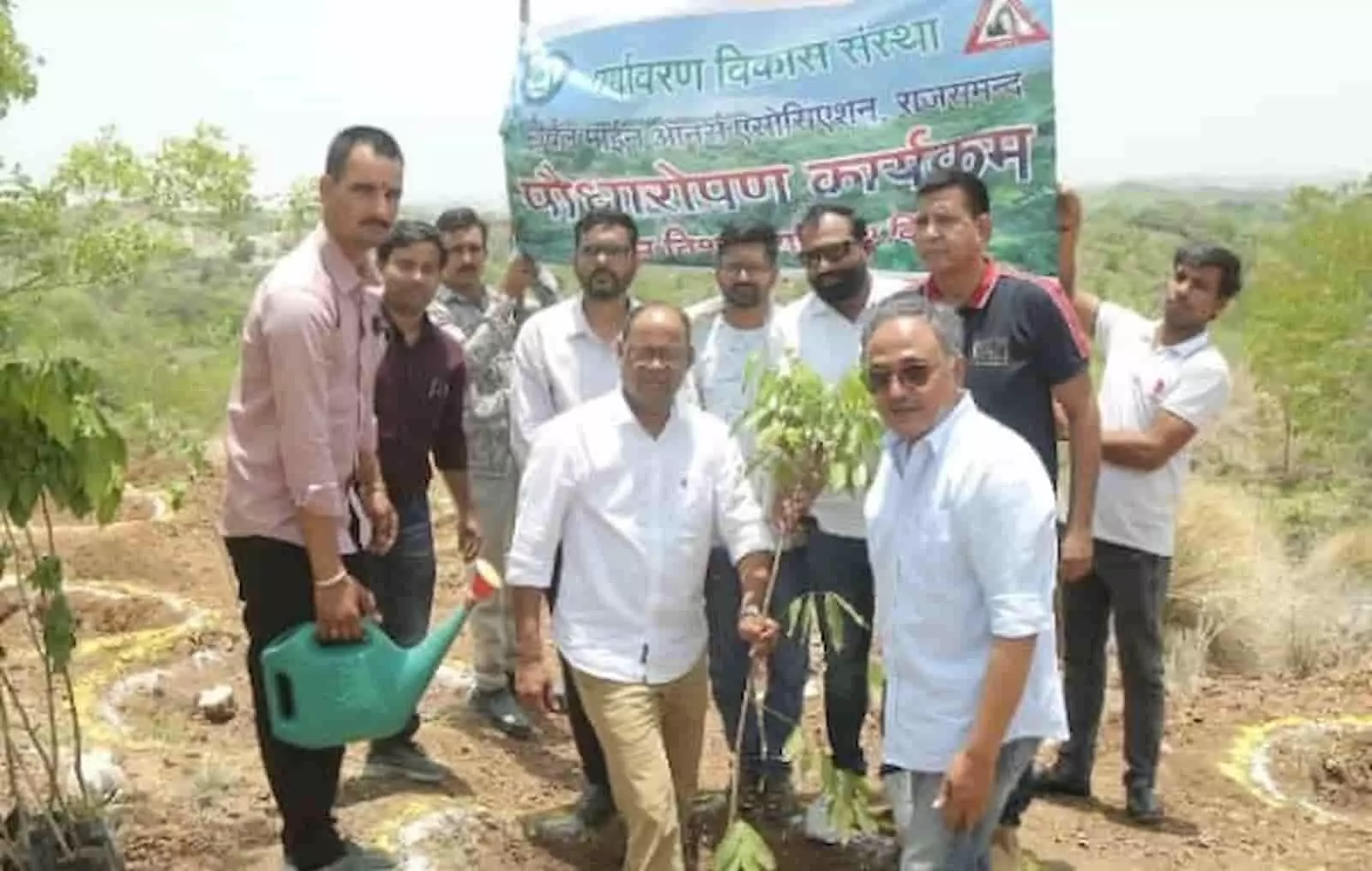 केलवा में बैनर के आगे पौधा पकड़े खड़े लोग पौधारोपण अभियान के संदर्भ में।
