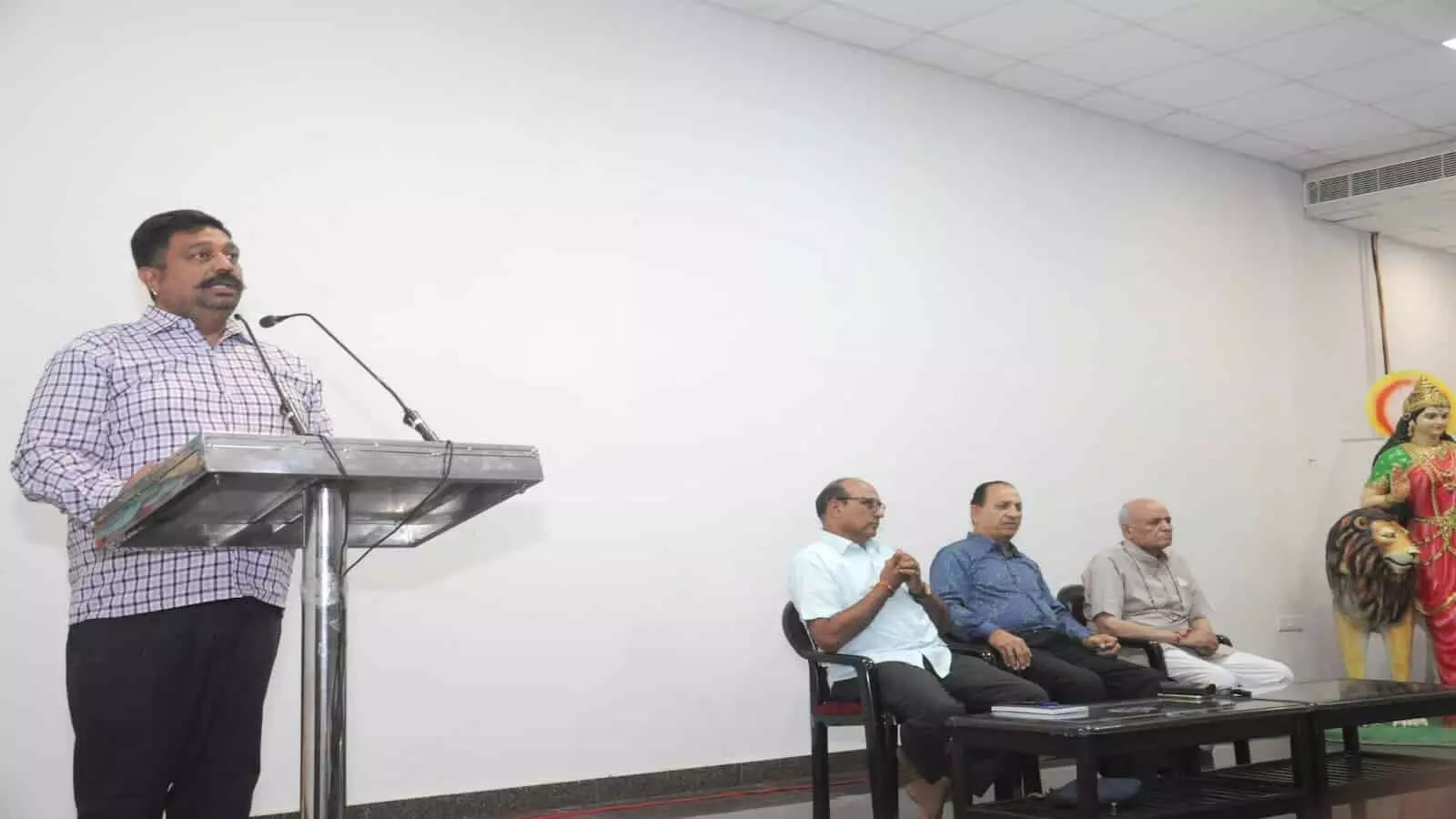 मंच पर पोडियम के सामने भाषण देते हुए व्यक्ति और कुर्सियों पर बैठे अन्य पदाधिकारी।