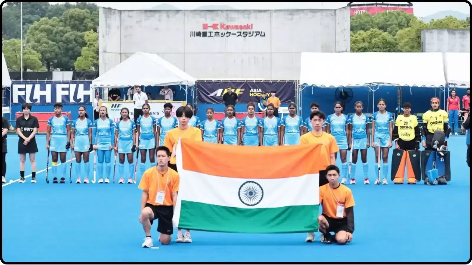 हॉकी मैदान पर तिरंगा थामे स्वयंसेवकों के पीछे कतार में खड़ी भारतीय महिला हॉकी टीम दिख रही है।