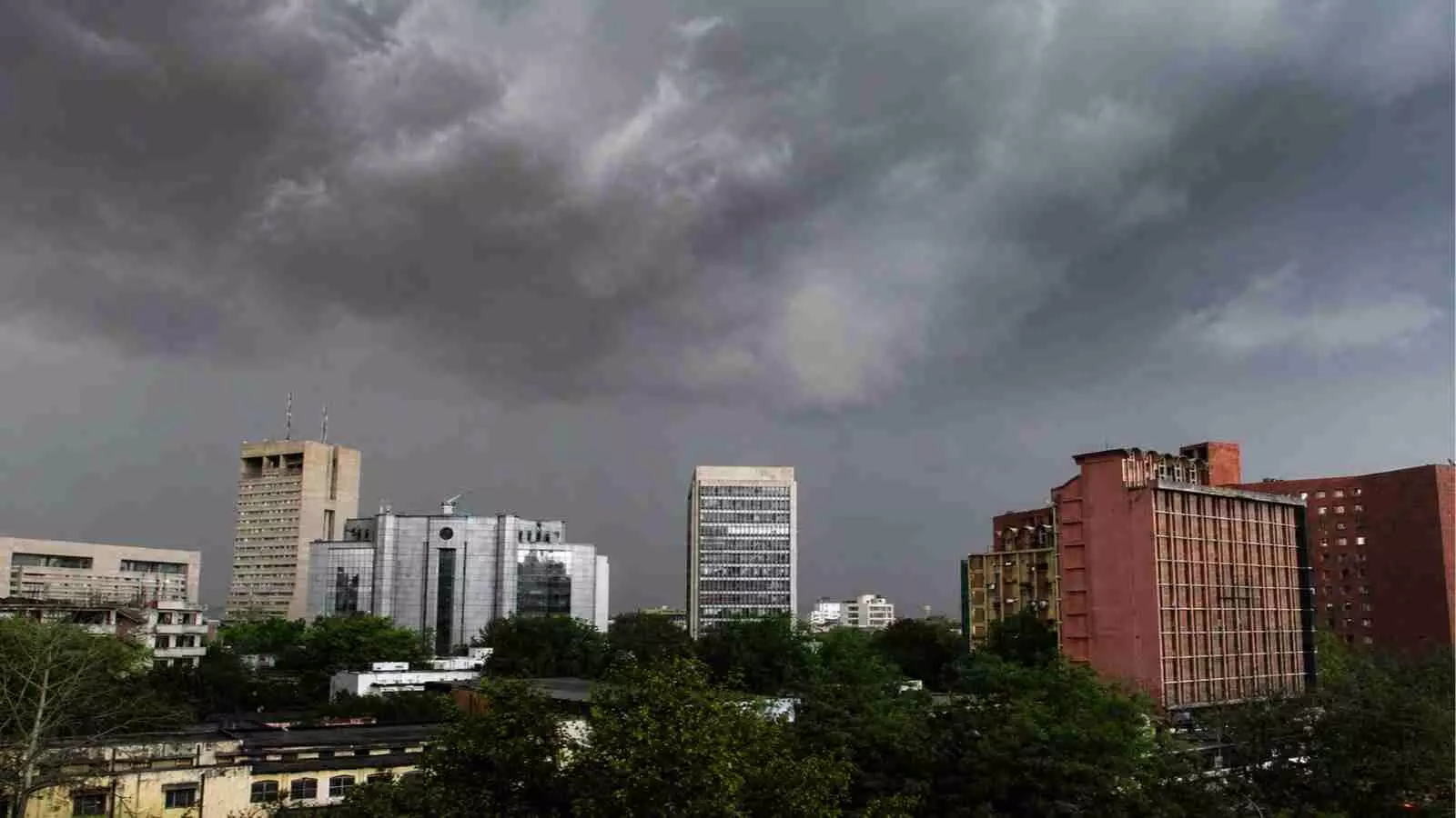 काले बादलों से घिरा हुआ आसमान और उसके नीचे बनी शहर की इमारतें दिखाई दे रही हैं।