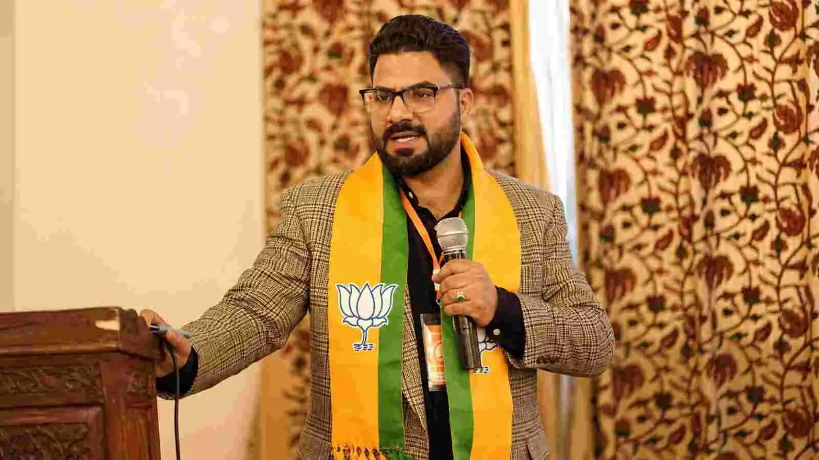 एक मंच पर खड़े होकर हाथ में माइक लिए भाषण देते हुए भाजपा नेता साजिद युसुफ शाह की तस्वीर।