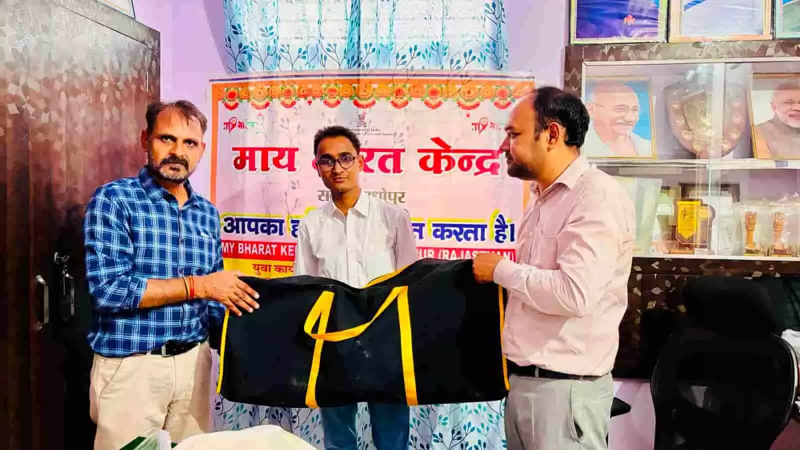 तस्वीर में तीन लोग माय भारत केंद्र के बैनर के सामने एक बड़ा काले और पीले रंग का खेल किट बैग पकड़े खड़े हैं। तस्वीर में तीन लोग माय भारत केंद्र के बैनर के सामने एक बड़ा काले और पीले रंग का खेल किट बैग पकड़े खड़े हैं।