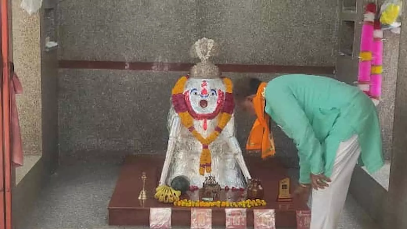 तस्वीर में एक व्यक्ति केसरिया पटका पहने हुए मंदिर के भीतर स्थापित हनुमान जी की प्रतिमा के सामने झुककर प्रणाम कर रहा है।