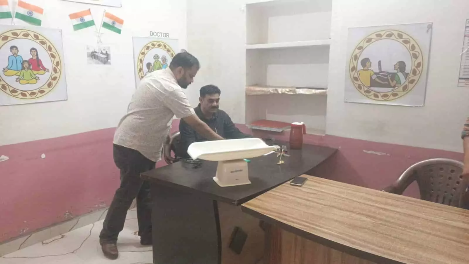 तस्वीर में चिकित्सा कक्ष के भीतर एक मेज के पास खड़े हुए दो व्यक्ति आयुष्मान आरोग्य मंदिर के निरीक्षण के संदर्भ में दिख रहे हैं। तस्वीर में चिकित्सा कक्ष के भीतर एक मेज के पास खड़े हुए दो व्यक्ति आयुष्मान आरोग्य मंदिर के निरीक्षण के संदर्भ में दिख रहे हैं।