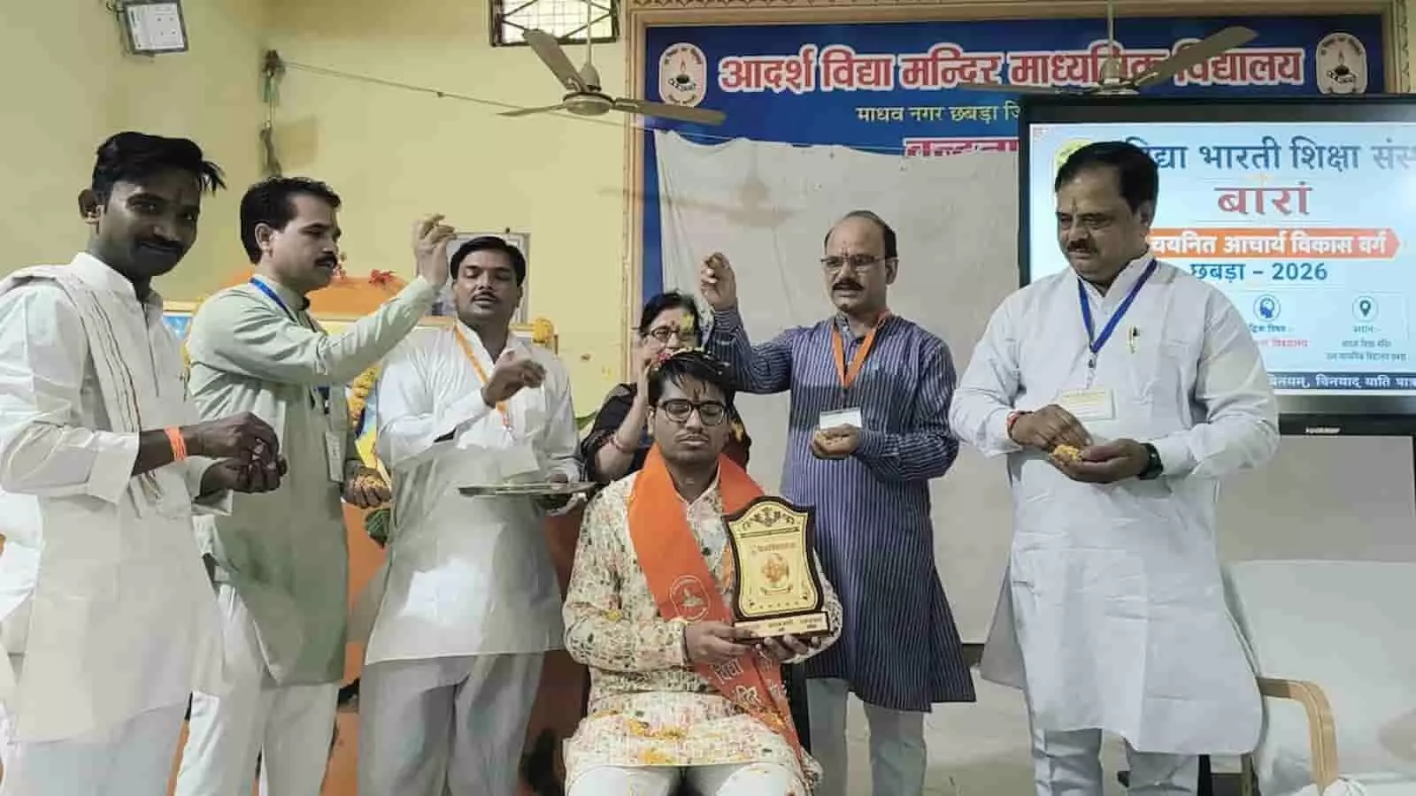 तस्वीर में एक मंच या हॉल के भीतर कई लोग खड़े होकर कुर्सी पर बैठे एक व्यक्ति पर फूल बरसाते हुए और उसे सम्मानित करते हुए दिख रहे हैं। तस्वीर में एक मंच या हॉल के भीतर कई लोग खड़े होकर कुर्सी पर बैठे एक व्यक्ति पर फूल बरसाते हुए और उसे सम्मानित करते हुए दिख रहे हैं।