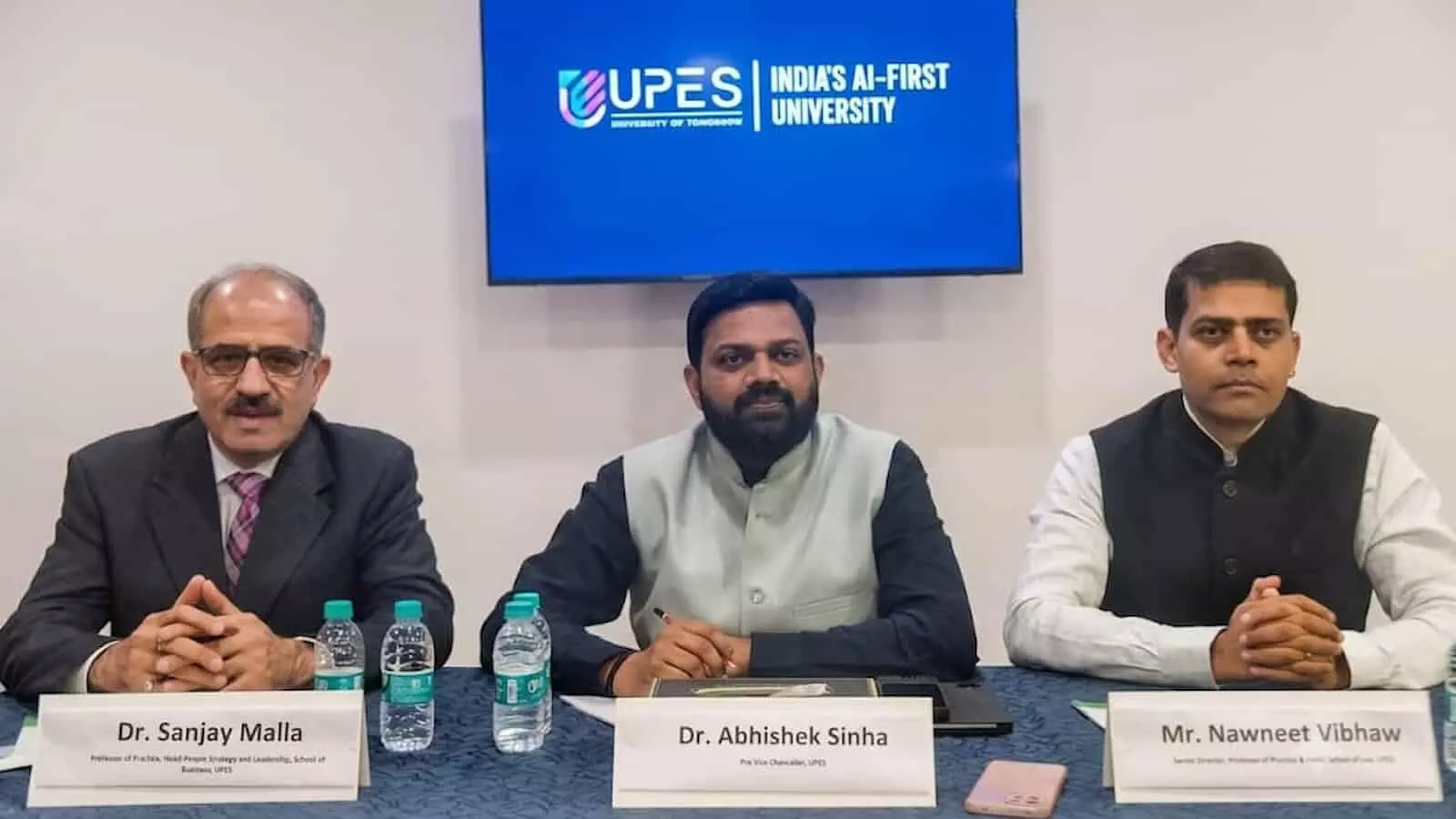 तस्वीर में पीछे UPES INDIAS AI-FIRST UNIVERSITY लिखे नीले डिजिटल स्क्रीन के सामने मेज पर नेमप्लेट और पानी की बोतलों के साथ बैठे तीन अधिकारी विश्वविद्यालय के नए विज़न के संदर्भ में दिख रहे हैं।