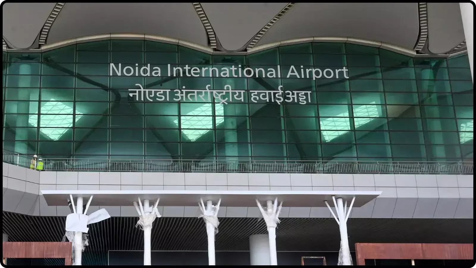 एयरपोर्ट की कांच वाली मुख्य इमारत के बाहर हिंदी और अंग्रेजी में नोएडा अंतर्राष्ट्रीय हवाई अड्डा लिखा हुआ जेवर में यात्री सेवाओं के संदर्भ में। एयरपोर्ट की कांच वाली मुख्य इमारत के बाहर हिंदी और अंग्रेजी में नोएडा अंतर्राष्ट्रीय हवाई अड्डा लिखा हुआ जेवर में यात्री सेवाओं के संदर्भ में।