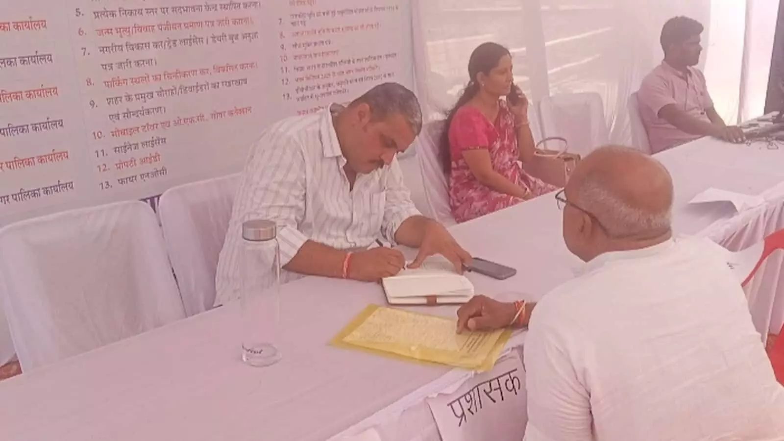 तस्वीर में एक शिविर के भीतर प्रशासनिक कर्मचारी मेज पर बैठकर कागजी कार्रवाई पूरी करते हुए और उनके सामने एक व्यक्ति बैठा हुआ दिख रहा है। तस्वीर में एक शिविर के भीतर प्रशासनिक कर्मचारी मेज पर बैठकर कागजी कार्रवाई पूरी करते हुए और उनके सामने एक व्यक्ति बैठा हुआ दिख रहा है।