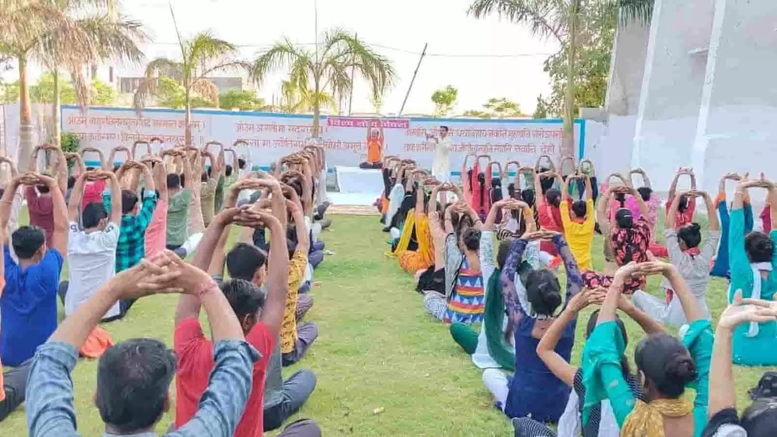 तस्वीर में कई लोग एक खुले मैदान में हरी घास पर बैठकर दोनों हाथ ऊपर उठाकर योग आसनों का सामूहिक अभ्यास कर रहे हैं।