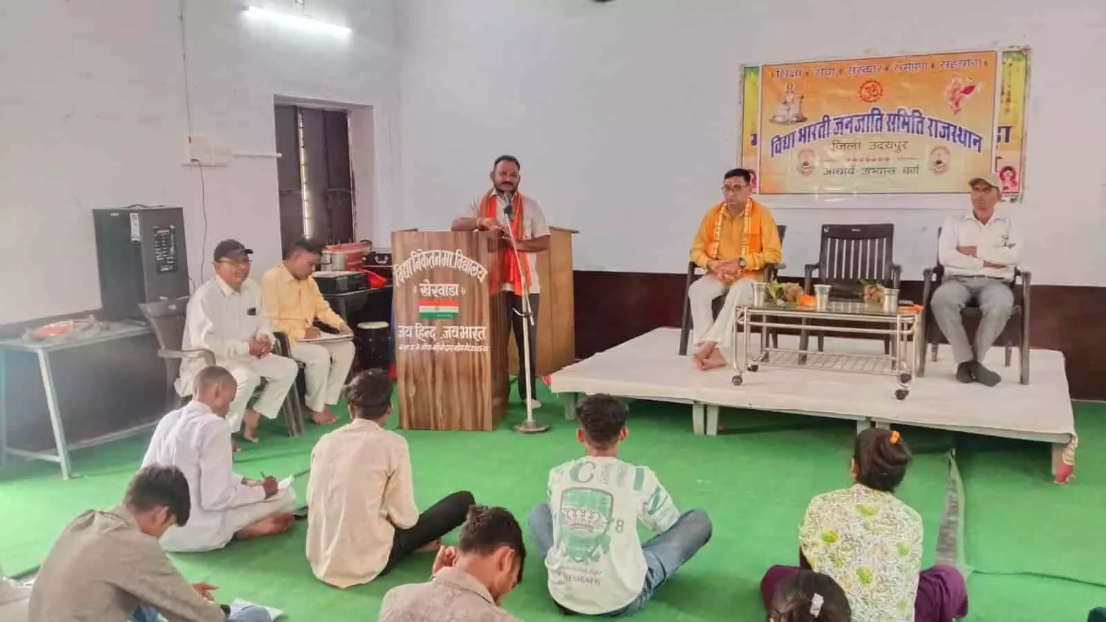 तस्वीर में एक बड़े हॉल के भीतर फर्श पर हरी चटाई पर बैठकर शिक्षार्थी मंच पर खड़े वक्ता का व्याख्यान सुन रहे हैं। तस्वीर में एक बड़े हॉल के भीतर फर्श पर हरी चटाई पर बैठकर शिक्षार्थी मंच पर खड़े वक्ता का व्याख्यान सुन रहे हैं।