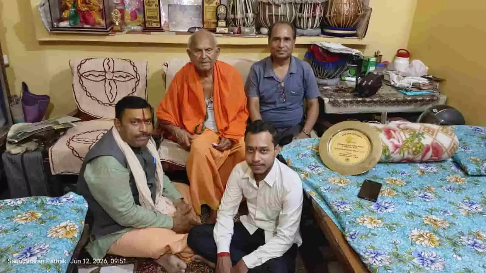 तस्वीर में एक कमरे के भीतर भगवा वस्त्र पहने बुजुर्ग संगीत गुरु पंडित परशुराम पाठक और अन्य लोग बैठे हुए दिख रहे हैं। तस्वीर में एक कमरे के भीतर भगवा वस्त्र पहने बुजुर्ग संगीत गुरु पंडित परशुराम पाठक और अन्य लोग बैठे हुए दिख रहे हैं।