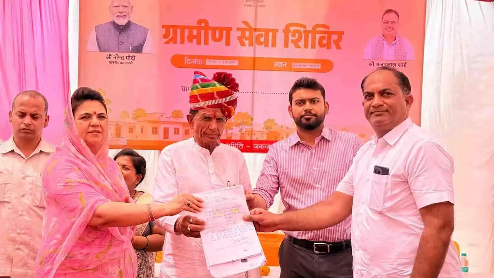 तस्वीर में पीछे ग्रामीण सेवा शिविर का बैनर लगा है, जिसके आगे राजसमंद सांसद महिमा कुमारी मेवाड़, पारंपरिक साफा पहने एक बुजुर्ग और अन्य अधिकारी एक दस्तावेज को हाथ में थामे हुए खड़े हैं। तस्वीर में पीछे ग्रामीण सेवा शिविर का बैनर लगा है, जिसके आगे राजसमंद सांसद महिमा कुमारी मेवाड़, पारंपरिक साफा पहने एक बुजुर्ग और अन्य अधिकारी एक दस्तावेज को हाथ में थामे हुए खड़े हैं।