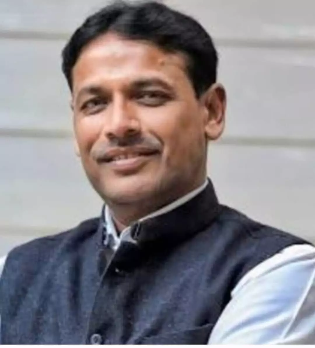 तस्वीर में मुस्कुराते हुए एक पुरुष का क्लोज-अप पोर्ट्रेट दिख रहा है, जिन्होंने सफेद शर्ट के ऊपर गहरे रंग की सदरी (नेहरू जैकेट) पहनी हुई है।