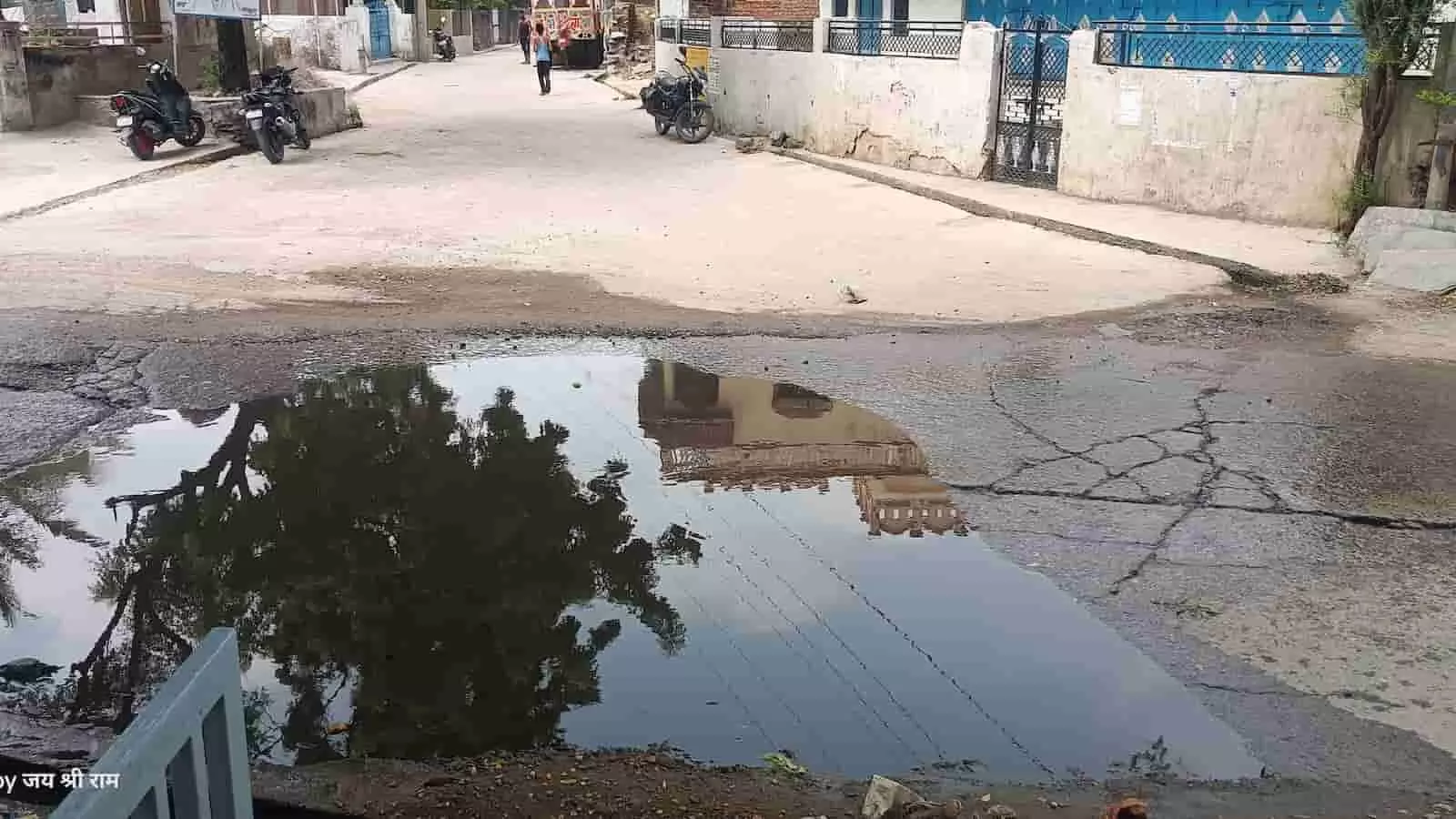 तस्वीर में एक पक्की ग्रामीण सड़क के बीचों-बीच एक बड़ा और गहरा गंदे पानी का गड्ढा (जलजमाव) दिखाई दे रहा है, जिसमें आसपास के मकानों और पेड़ों का प्रतिबिंब साफ नजर आ रहा है।