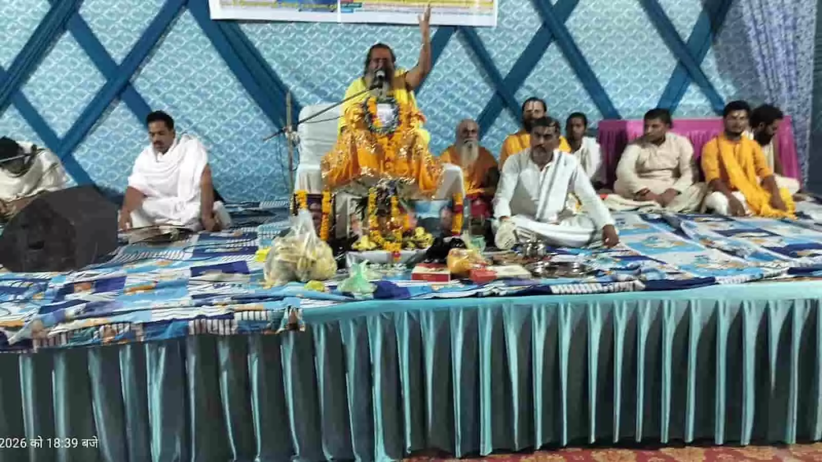 तस्वीर में एक बड़े धार्मिक मंच पर सुंदर वस्त्रों और मालाओं से सुसज्जित गद्दी पर एक कथावाचक महाराज हाथ उठाकर प्रवचन दे रहे हैं तथा उनके आस-पास अन्य संत व सेवादार बैठे हुए हैं।