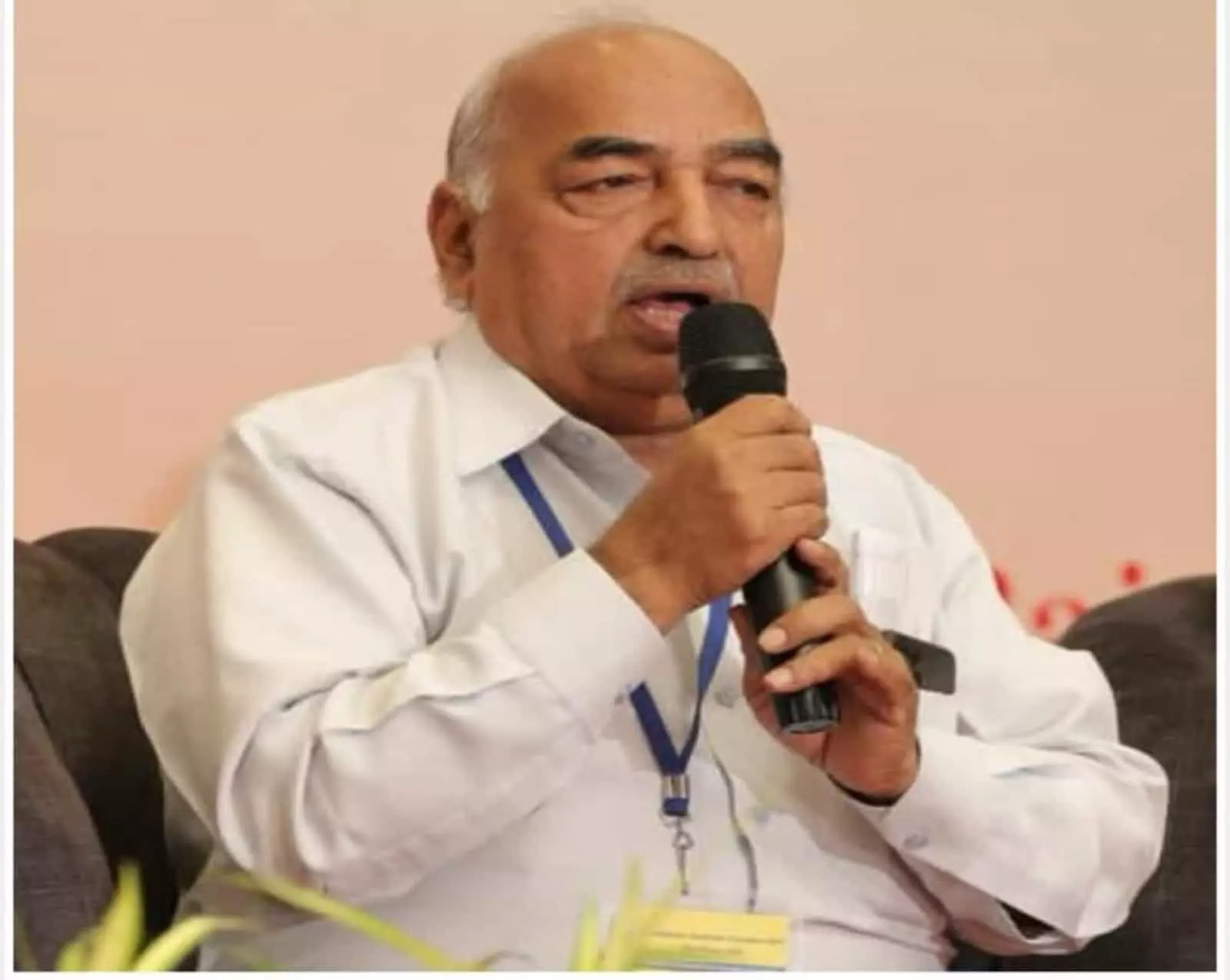 तस्वीर में एक वरिष्ठ प्रबुद्ध वक्ता सोफे पर बैठे हुए हैं, जो हाथ में काला माइक थामे हुए अत्यंत गंभीर मुद्रा में संबोधित कर रहे हैं।