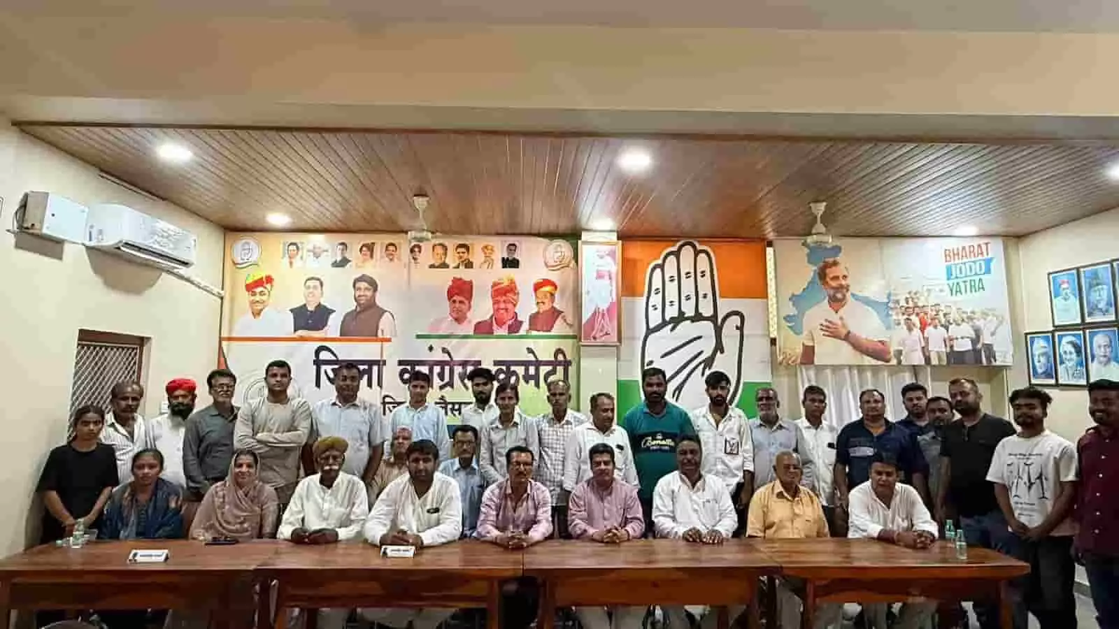 तस्वीर में एक बड़े हॉल के भीतर लंबी मेज के पीछे कई कांग्रेस कार्यकर्ता और पदाधिकारी बैठे हुए हैं