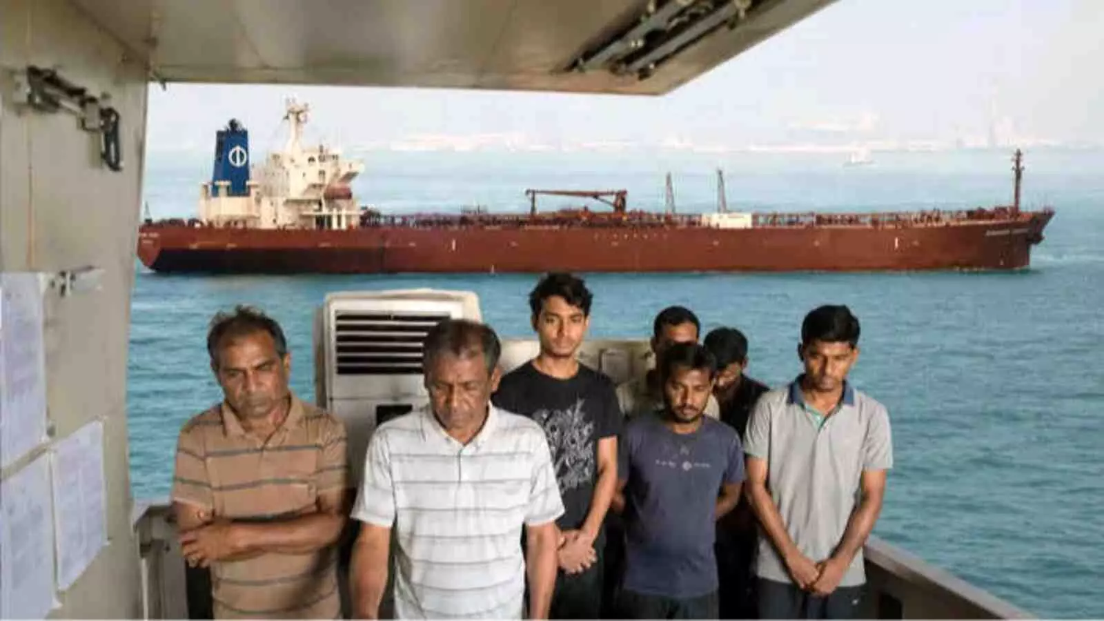 समुद्र के बीच खड़े एक जहाज की डेक पर खड़े छह उदास चालक दल के सदस्य और उनके पीछे तैरता बड़ा मालवाहक जहाज| समुद्र के बीच खड़े एक जहाज की डेक पर खड़े छह उदास चालक दल के सदस्य और उनके पीछे तैरता बड़ा मालवाहक जहाज|