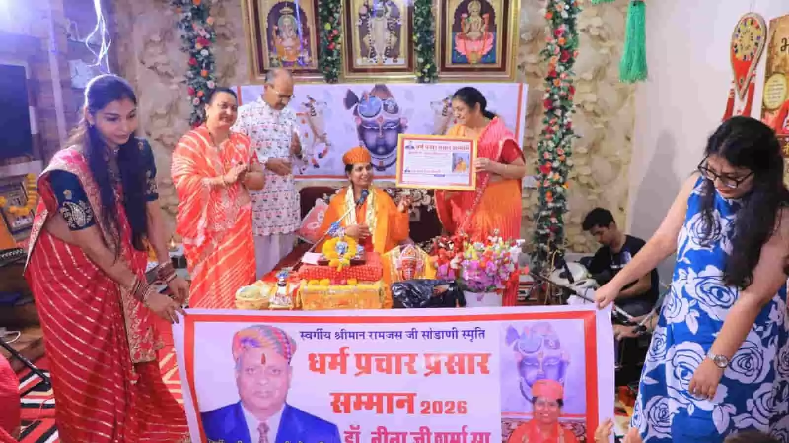 धार्मिक समारोह में बैनर के पीछे खड़े अतिथि डॉ. नीना शर्मा को स्मृति चिन्ह और अभिनंदन पत्र सौंपकर सम्मानित कर रहे हैं।