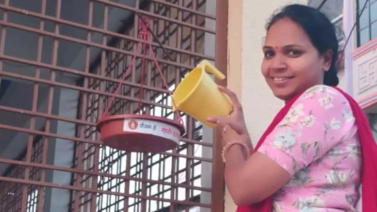 लोहे की जाली पर लटके लाल रंग के परिंडे में पीले मग से पानी डालती महिला। लोहे की जाली पर लटके लाल रंग के परिंडे में पीले मग से पानी डालती महिला।