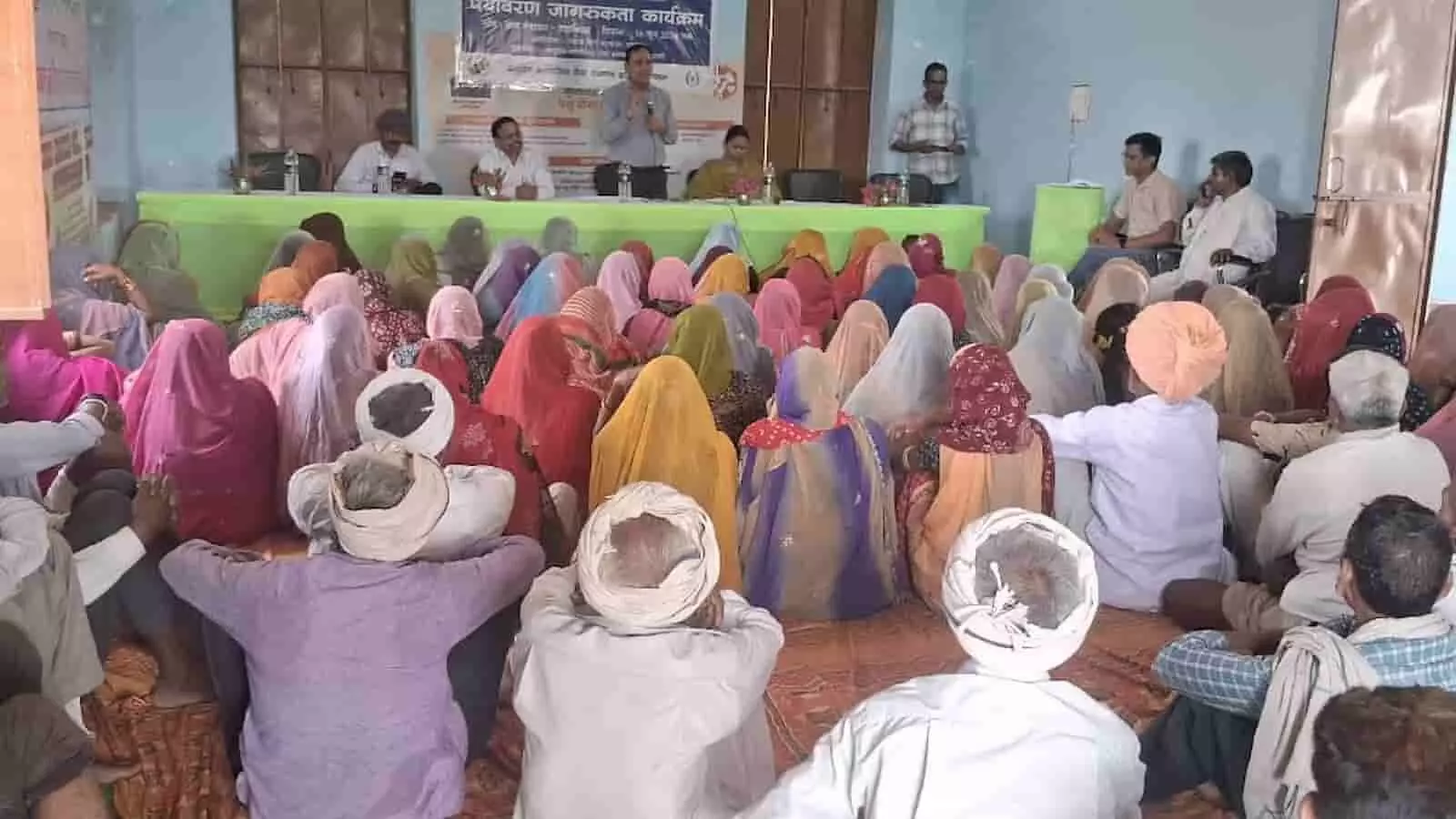 टटाकिया ग्राम पंचायत के सामुदायिक भवन में पर्यावरण जागरूकता कार्यक्रम के दौरान मंच पर बैठे अतिथि और सामने जमीन पर बैठे ग्रामीण नागरिक।