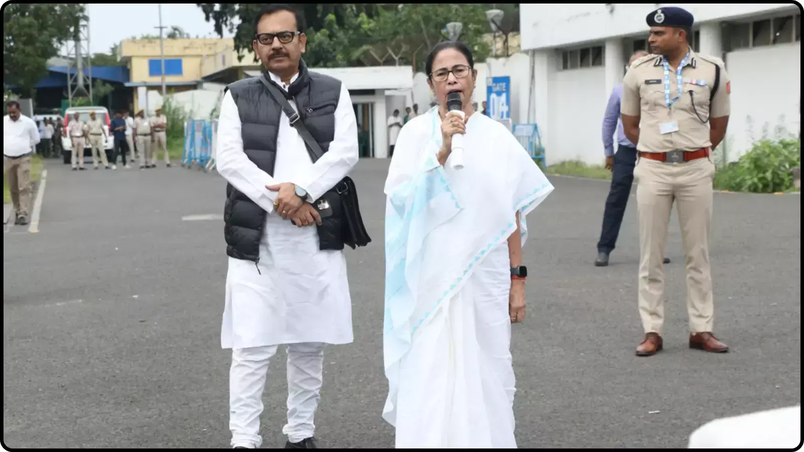 माइक थामकर बोलती महिला और बगल में खड़ा पुरुष सुरक्षाकर्मियों के साथ बाहरी परिसर में राजनीतिक विमर्श के संदर्भ में दिख रहे हैं। माइक थामकर बोलती महिला और बगल में खड़ा पुरुष सुरक्षाकर्मियों के साथ बाहरी परिसर में राजनीतिक विमर्श के संदर्भ में दिख रहे हैं।