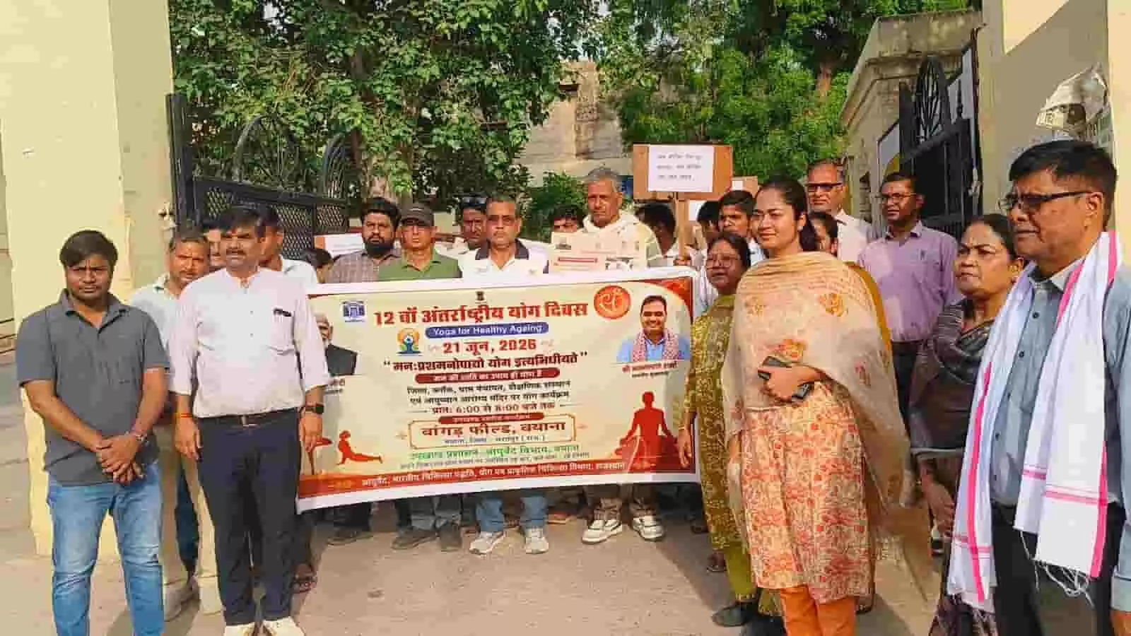 बयाना में अंतर्राष्ट्रीय योग दिवस के बैनर के साथ जागरूकता रैली में खड़े सरकारी कर्मचारी और नागरिक। बयाना में अंतर्राष्ट्रीय योग दिवस के बैनर के साथ जागरूकता रैली में खड़े सरकारी कर्मचारी और नागरिक।