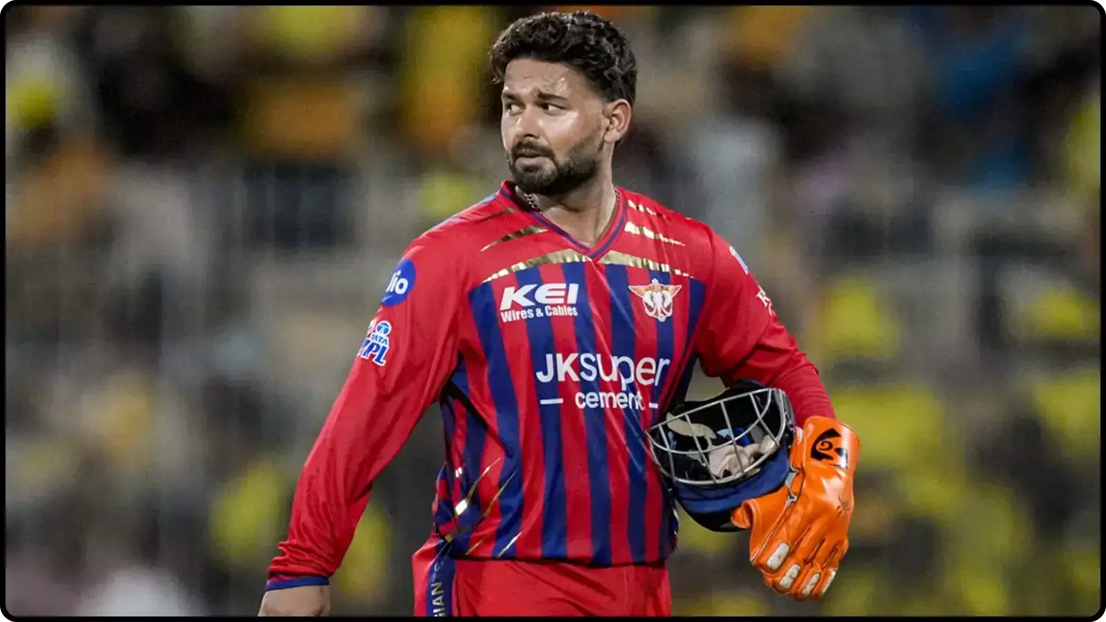 क्रिकेट मैदान पर लाल और नीली जर्सी पहने रिषभ पंत हाथ में हेलमेट लिए दिख रहे हैं।