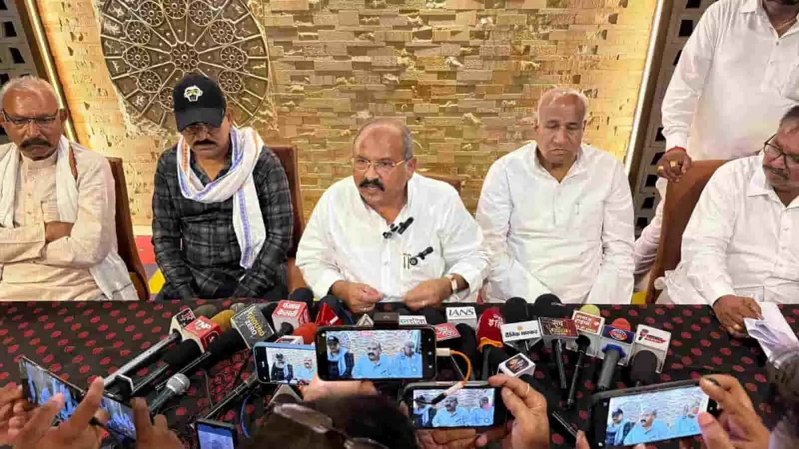 कोटा में आयोजित प्रेस वार्ता के दौरान टेबल पर रखे मीडिया माइक के सामने बोलते पूर्व विधायक प्रहलाद गुंजल और अन्य नेता।