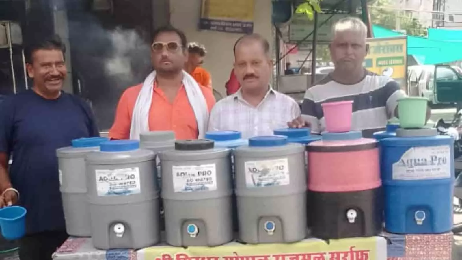 मेज पर रखे कई नीले और ग्रे रंग के कैंपरों के पीछे खड़े चार पुरुष। मेज पर रखे कई नीले और ग्रे रंग के कैंपरों के पीछे खड़े चार पुरुष।