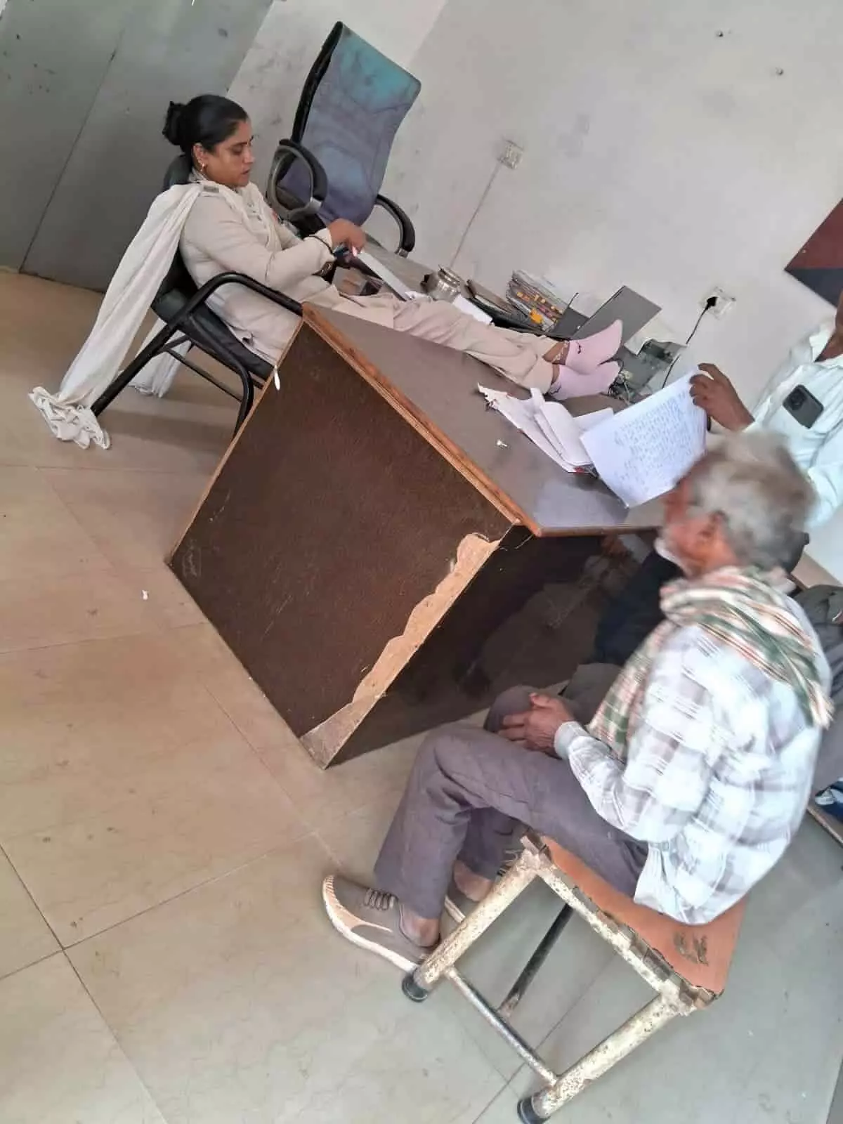 मेज पर पैर रखकर बैठी महिला पुलिसकर्मी और सामने बैठा एक बुजुर्ग व्यक्ति। मेज पर पैर रखकर बैठी महिला पुलिसकर्मी और सामने बैठा एक बुजुर्ग व्यक्ति।