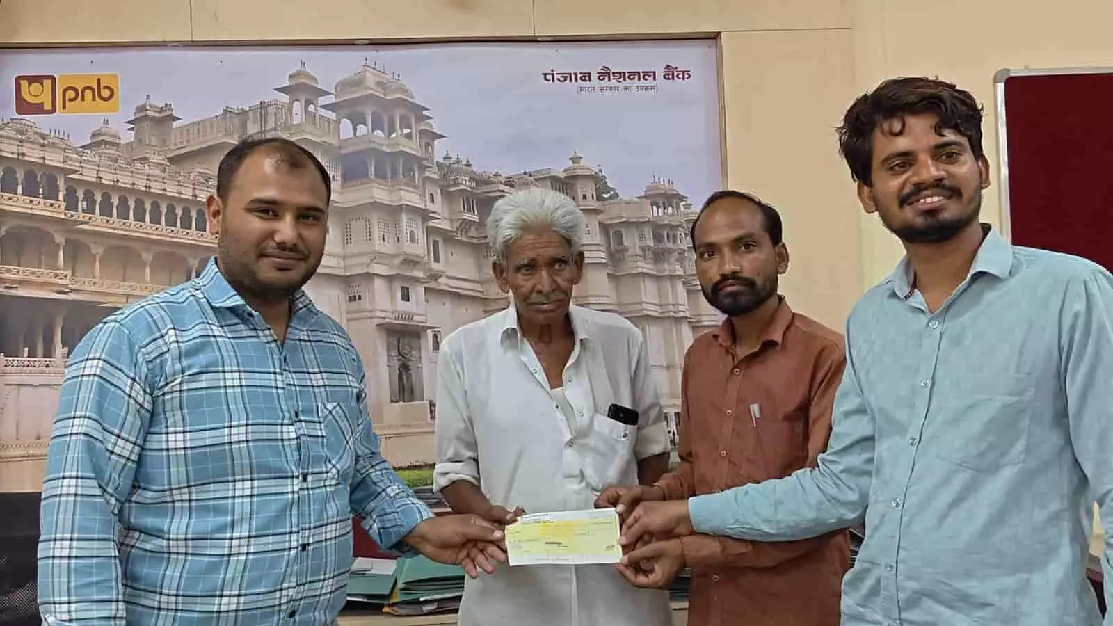पंजाब नेशनल बैंक में चार पुरुष एक बुजुर्ग व्यक्ति को बैंक चेक प्रदान करते हुए।