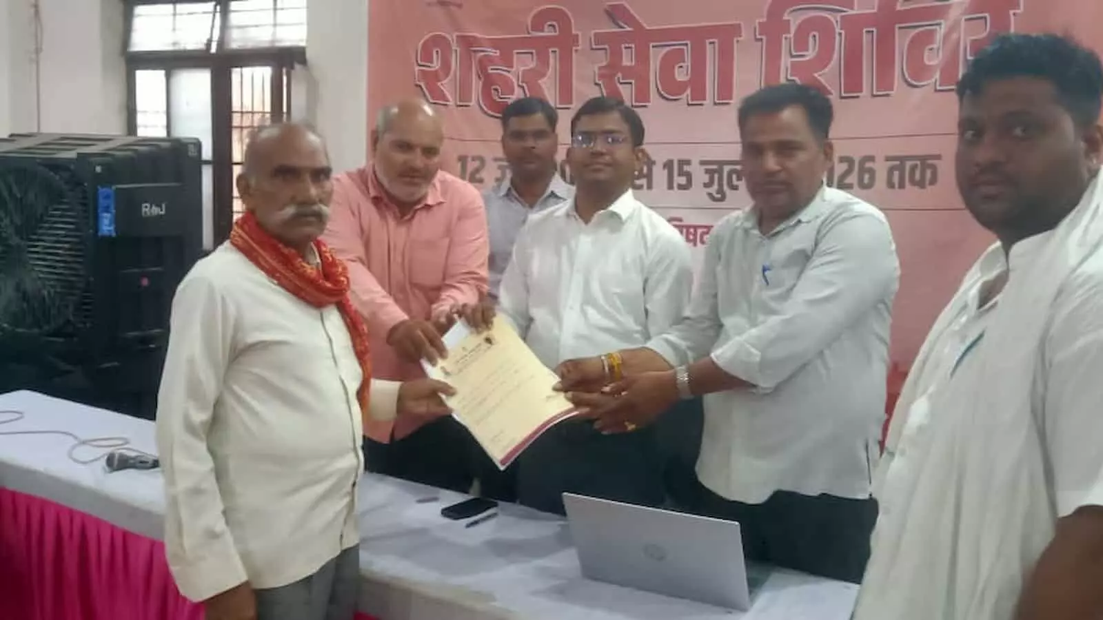 शहरी सेवा शिविर में अधिकारियों द्वारा बुजुर्ग व्यक्ति को प्रमाण पत्र सौंपने का दृश्य। शहरी सेवा शिविर में अधिकारियों द्वारा बुजुर्ग व्यक्ति को प्रमाण पत्र सौंपने का दृश्य।