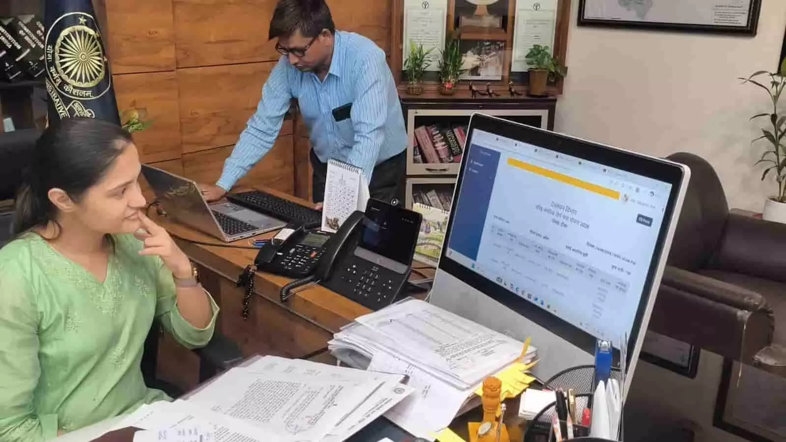 कार्यालय में कम्प्यूटर स्क्रीन और दस्तावेजों के साथ वरिष्ठ नागरिक तीर्थ यात्रा योजना की लॉटरी निकालते अधिकारी।