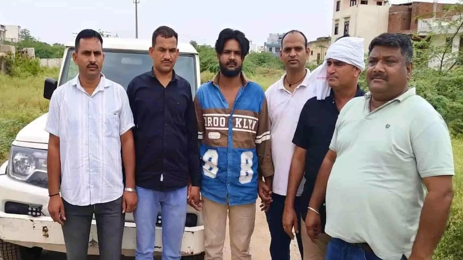 एक सफेद वाहन के आगे खड़े पुलिसकर्मी और हिरासत में लिया गया व्यक्ति। एक सफेद वाहन के आगे खड़े पुलिसकर्मी और हिरासत में लिया गया व्यक्ति।