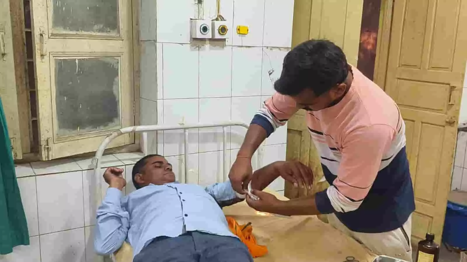 अस्पताल के बिस्तर पर लेटे घायल युवक के हाथ पर पट्टी बांधी जा रही है।