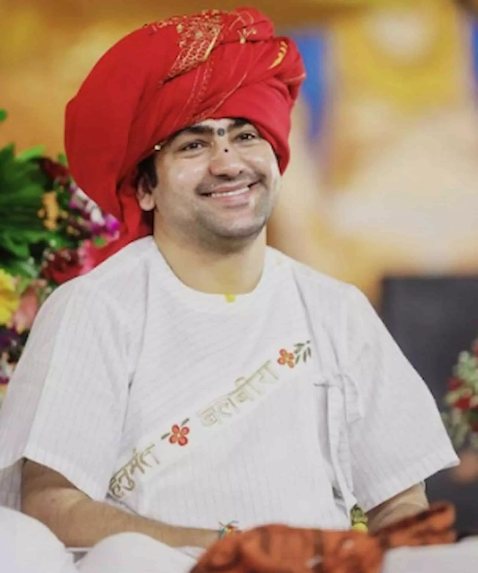तस्वीर में मुस्कुराते हुए बागेश्वर धाम सरकार धीरेंद्र कृष्ण शास्त्री पारंपरिक वेशभूषा में बैठे हैं।