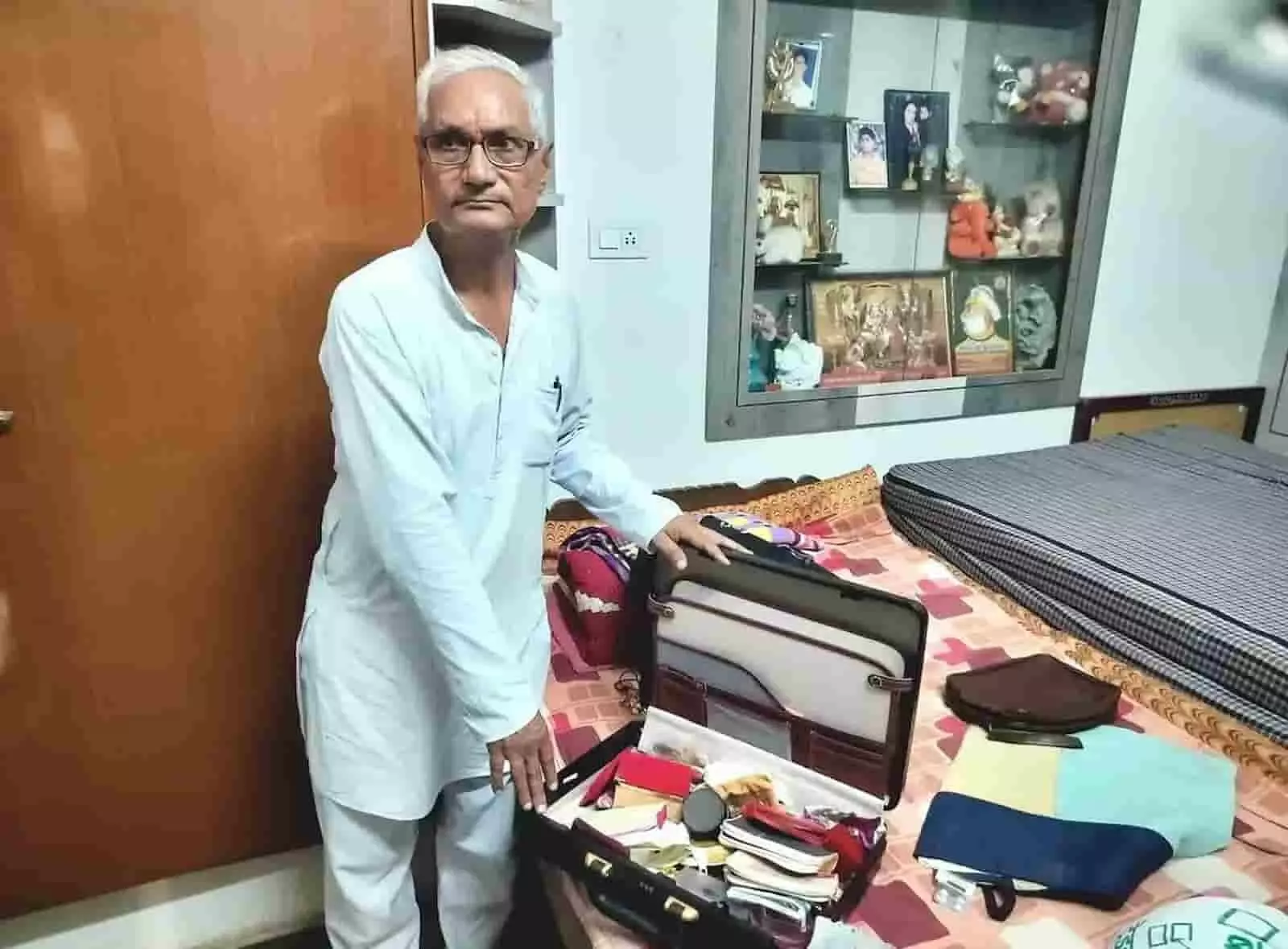 तस्वीर में कमरे के अंदर बेड पर खुले सूटकेस के पास खड़े बुजुर्ग व्यक्ति दिख रहे हैं। तस्वीर में कमरे के अंदर बेड पर खुले सूटकेस के पास खड़े बुजुर्ग व्यक्ति दिख रहे हैं।