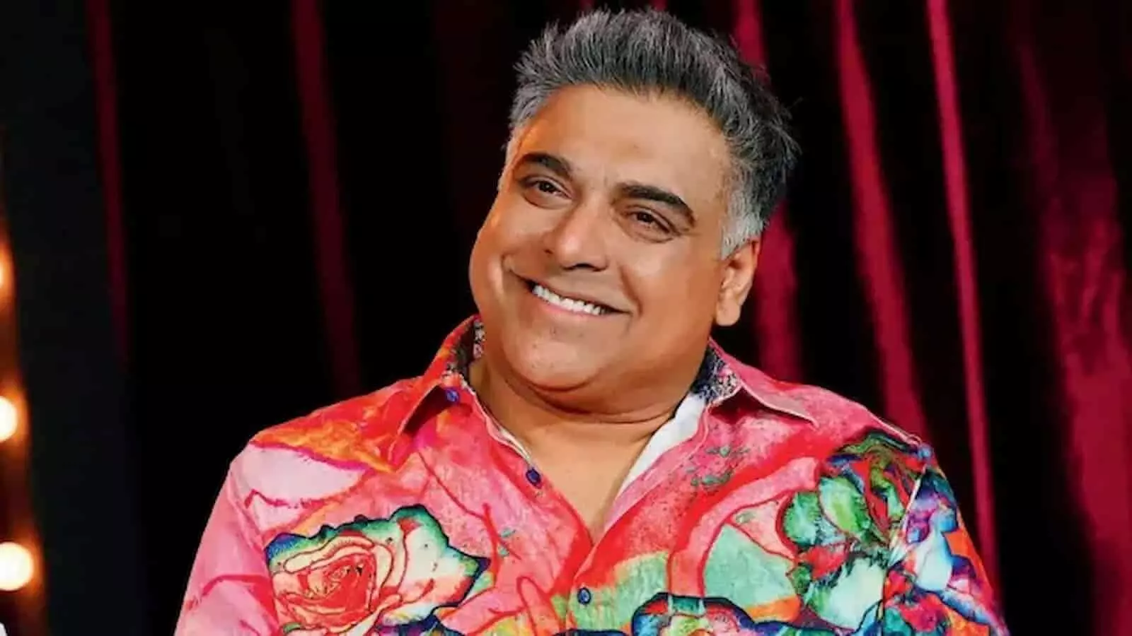 मुस्कुराते हुए अभिनेता राम कपूर गहरे लाल रंग के बैकग्राउंड के सामने खड़े हैं। मुस्कुराते हुए अभिनेता राम कपूर गहरे लाल रंग के बैकग्राउंड के सामने खड़े हैं।