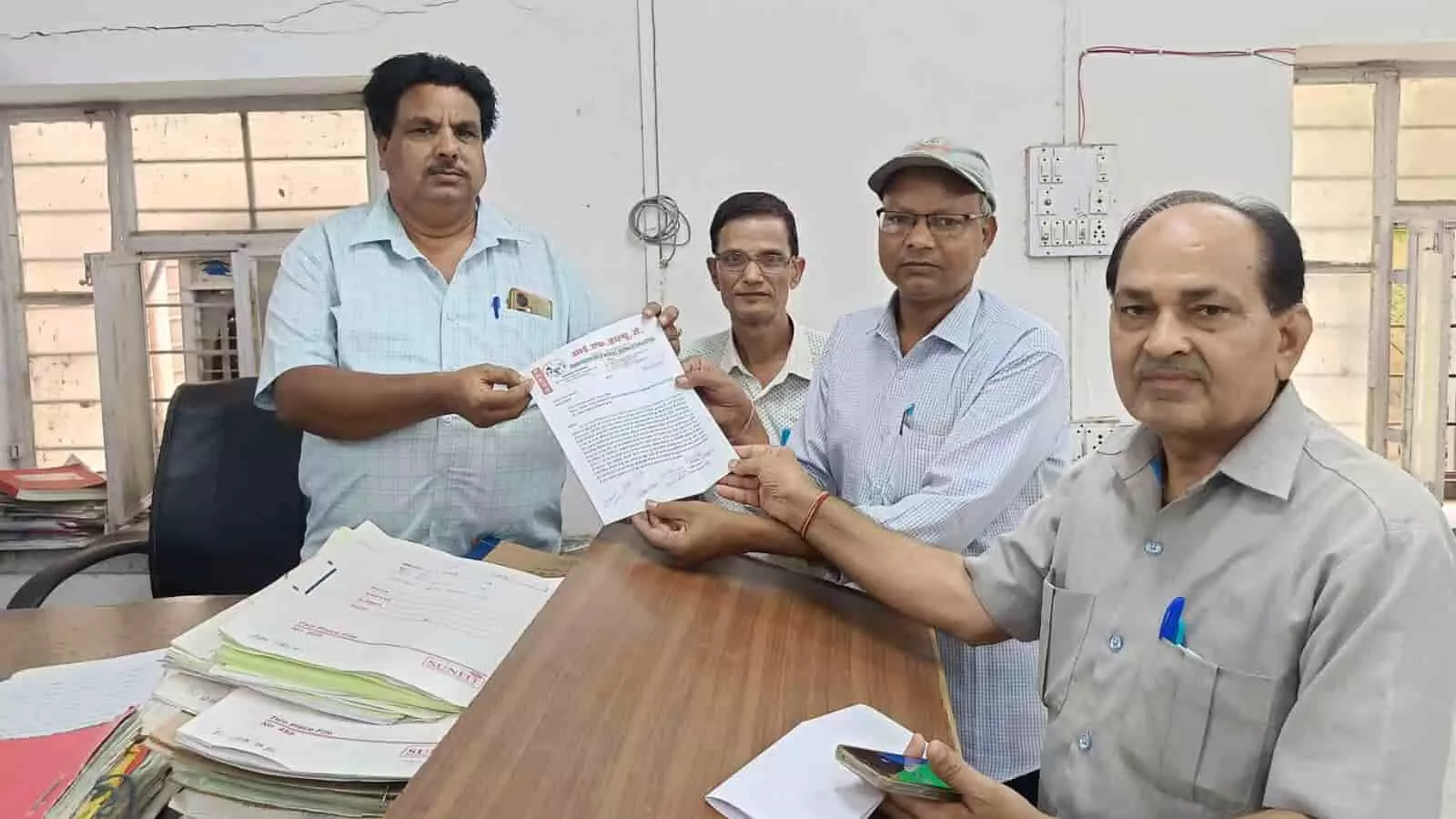 तस्वीर में एक कार्यालय के भीतर चार पुरुष एक अधिकारी को पत्र सौंपते हुए दिखाई दे रहे हैं।