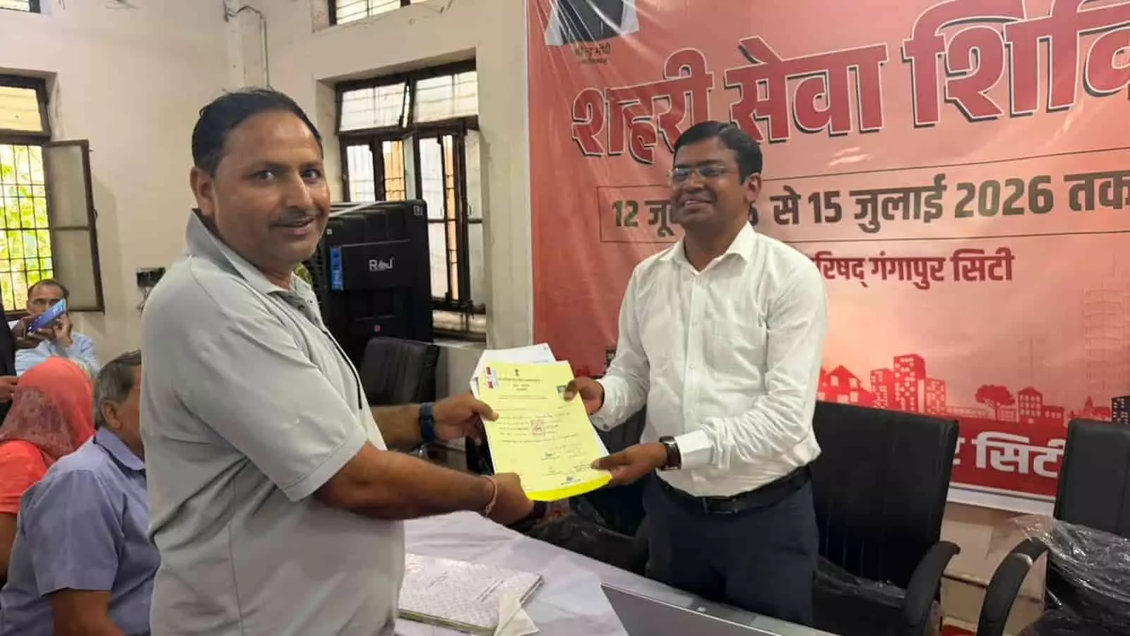 गंगापुर सिटी शहरी सेवा शिविर में अधिकारी नागरिक को सरकारी दस्तावेज सौंप रहे हैं।