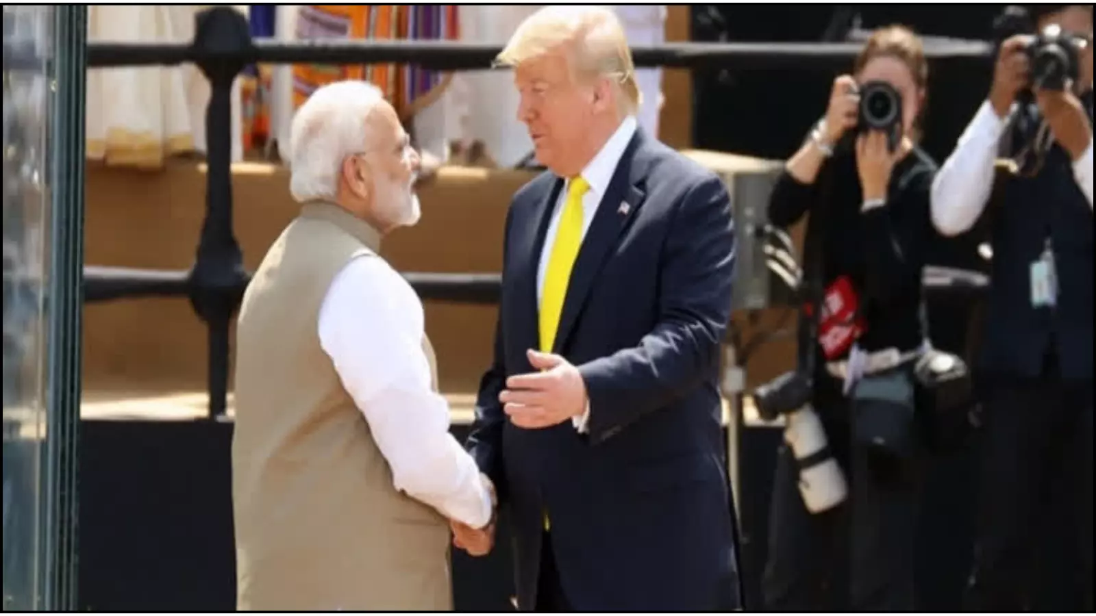 मंच पर हाथ मिलाते प्रधानमंत्री नरेंद्र मोदी और राष्ट्रपति डोनाल्ड ट्रंप पृष्ठभूमि में फोटोग्राफरों के सामने खड़े हैं।