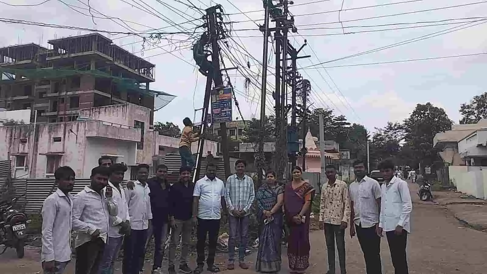 तस्वीर में बिजली के खंभे पर काम करते कर्मचारियों के पास खड़े लोग दिख रहे हैं।