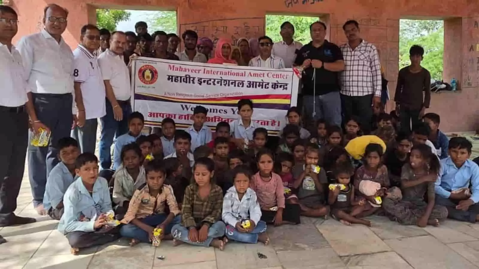 तस्वीर में महावीर इंटरनेशनल संस्था के सदस्य और बच्चे आमेट की भील बस्ती में बैनर के साथ बैठे हैं।