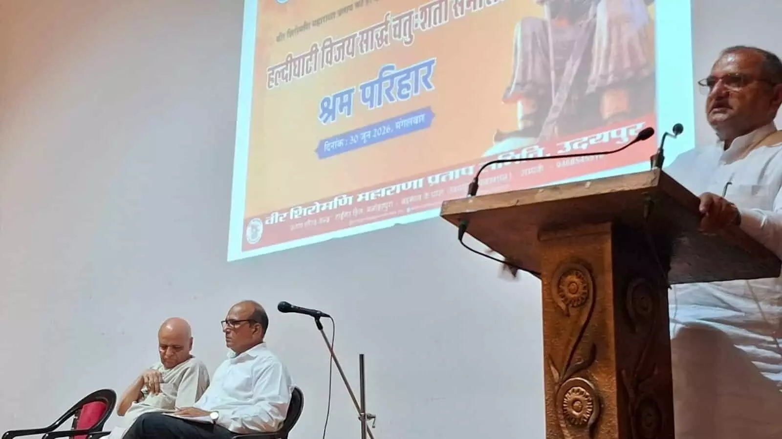 तस्वीर में कुंभा सभागार के मंच पर पोडियम से भाषण देते हुए निम्बाराम और बैठे हुए अन्य लोग दिख रहे हैं।