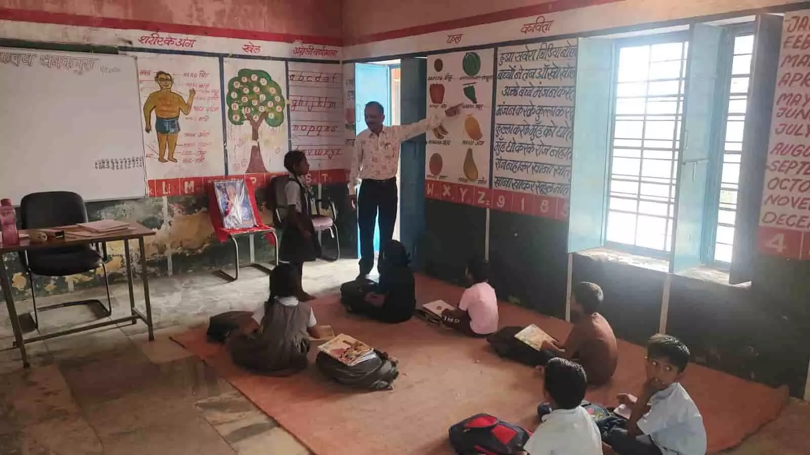 कक्षा के भीतर दीवार पर बने चार्ट की ओर इशारा करते शिक्षा अधिकारी और पढ़ाई करते छात्र।