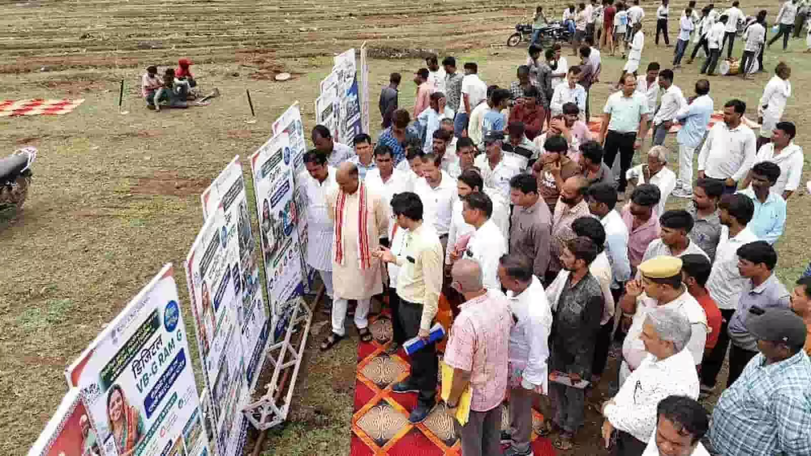 तस्वीर में खुले मैदान में अधिकारियों और ग्रामीणों की भीड़ योजना के बैनर देखते हुए। तस्वीर में खुले मैदान में अधिकारियों और ग्रामीणों की भीड़ योजना के बैनर देखते हुए।