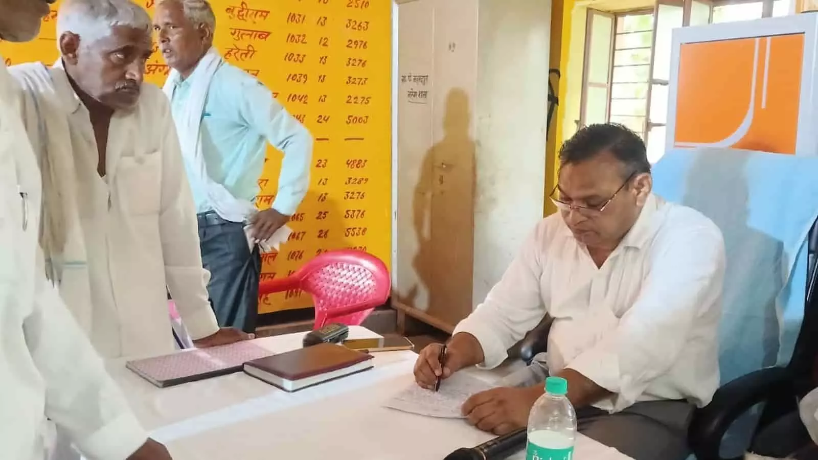 कार्यालय में मेज पर बैठकर लिखते हुए अधिकारी और उनके सामने खड़े ग्रामीण। कार्यालय में मेज पर बैठकर लिखते हुए अधिकारी और उनके सामने खड़े ग्रामीण।