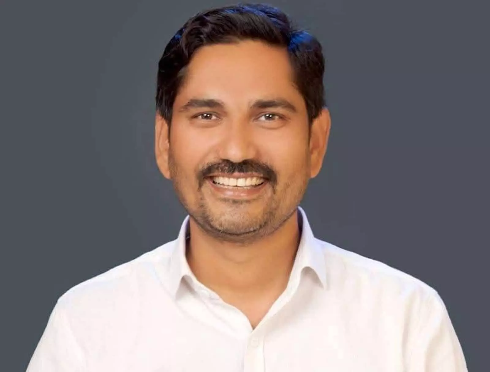 सफेद शर्ट पहने नरेंद्र मेघवाल एक गहरे रंग के बैकग्राउंड के सामने मुस्कुरा रहे हैं।