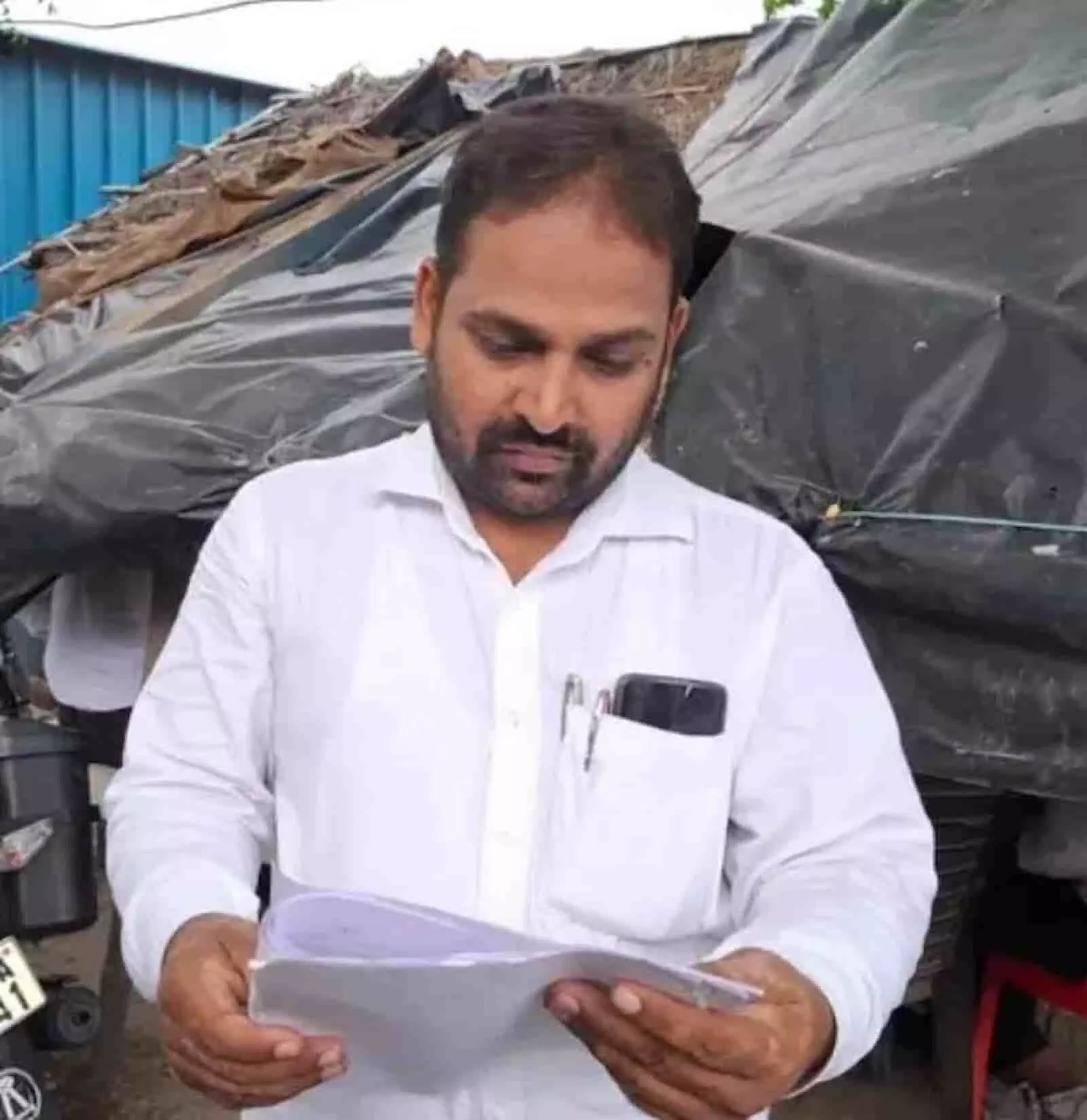 तस्वीर में सफेद शर्ट पहने एक व्यक्ति हाथ में दस्तावेज लिए हुए खड़ा दिखाई दे रहा है।